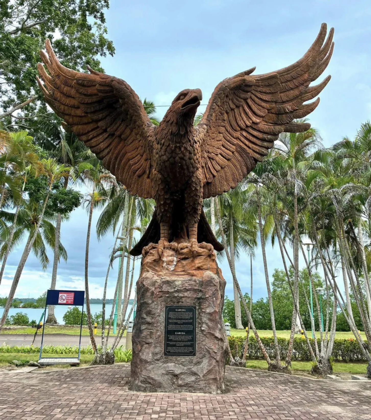Large carved wooden Garuda eagle statue with outstretched wings on a stone plinth, framed by palm trees on Bintan Island, Indonesia.
