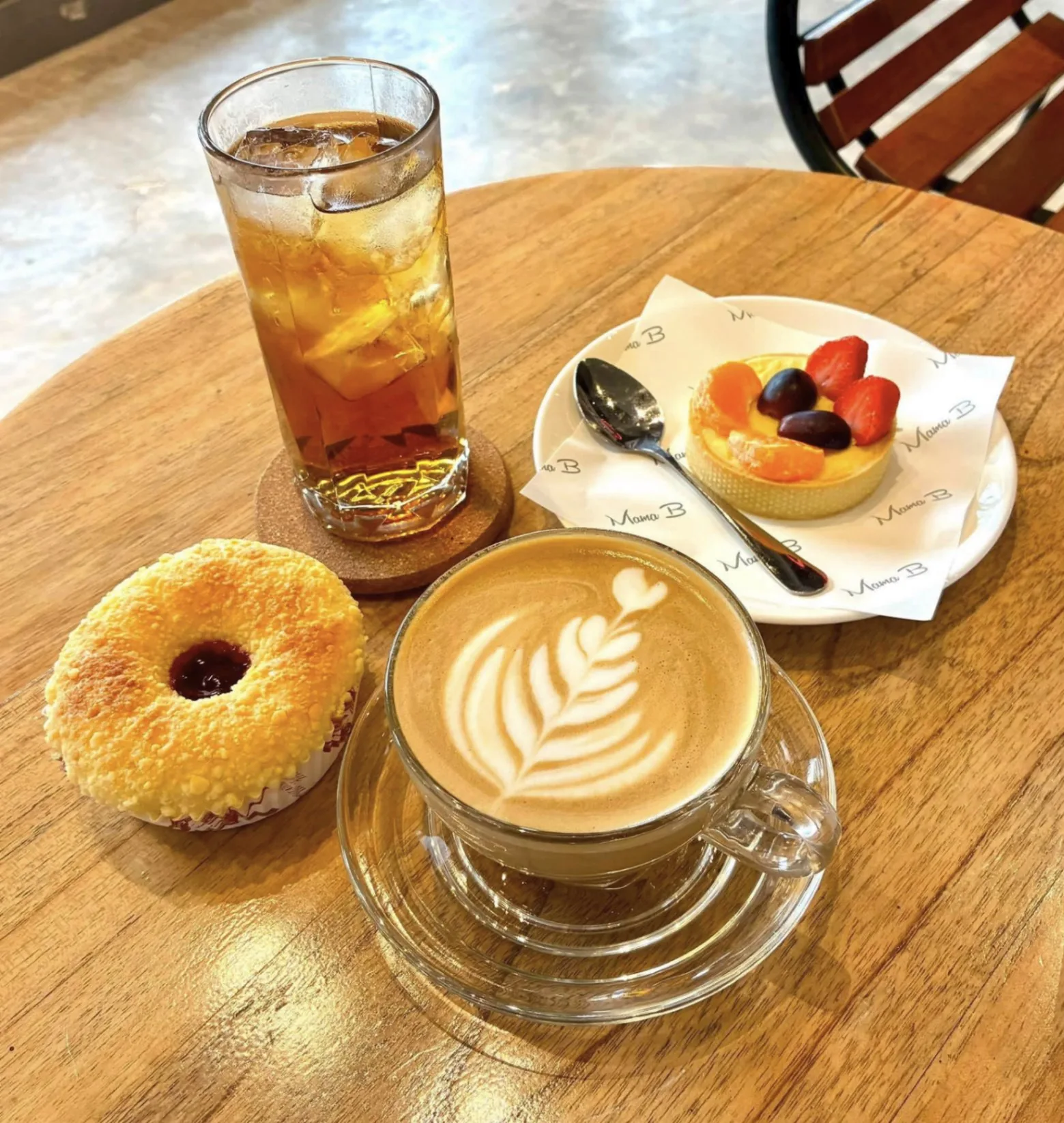 Cafe table with a glass of iced tea, a latte with leaf art, a jam-filled donut, and a fresh fruit tart at Mama B on Bintan Island, Indonesia.