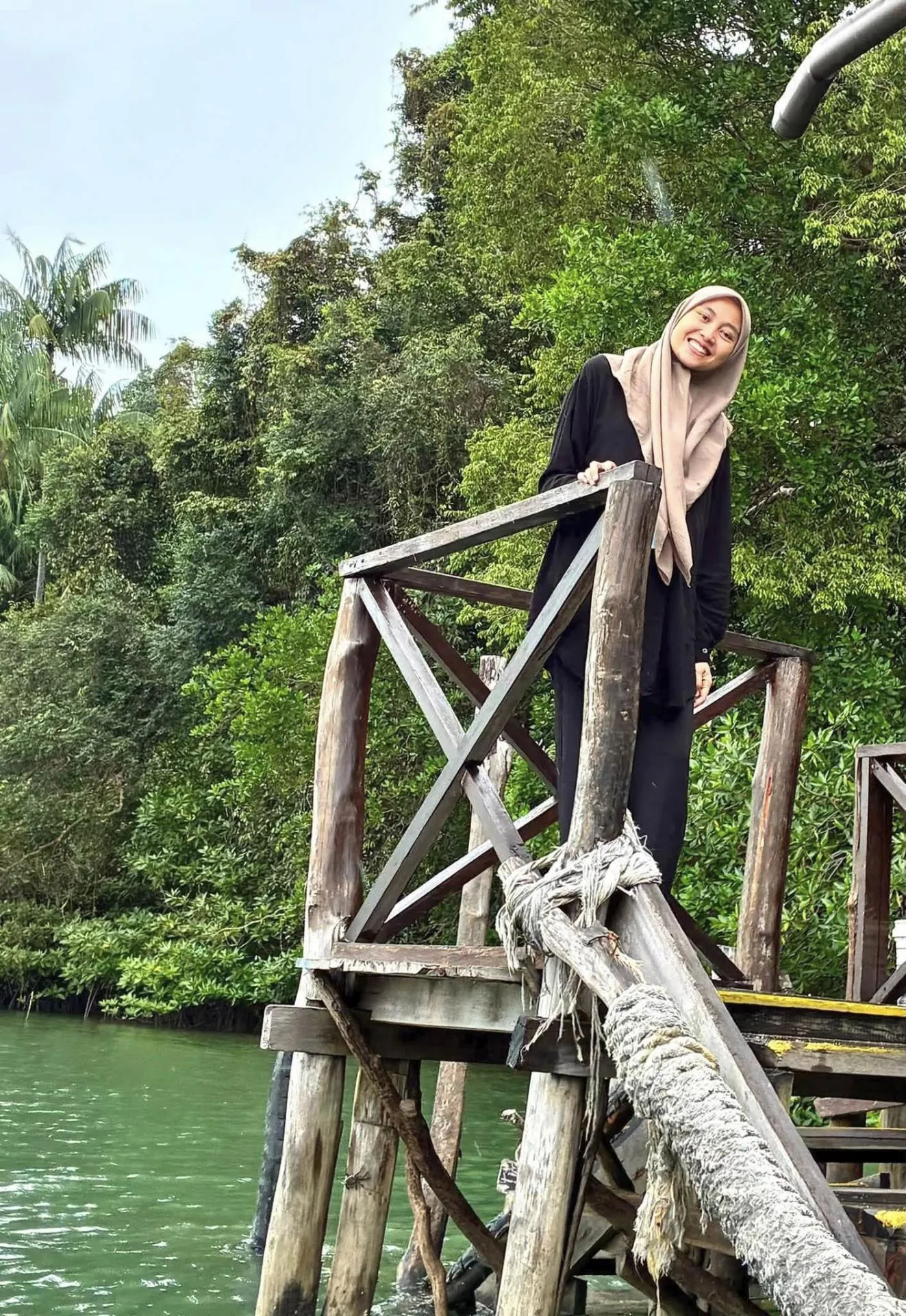 Smiling woman in a black outfit and blush hijab standing on a rustic wooden jetty above green mangrove water on Bintan Island, Indonesia.