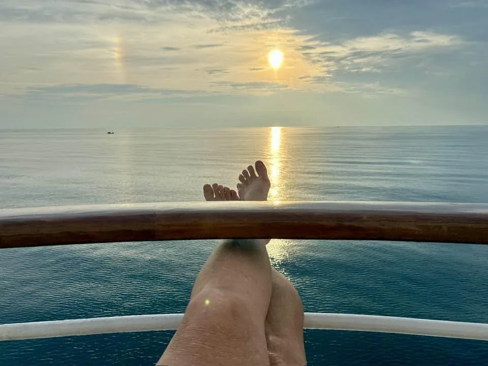 View of an ocean sunset from a ship's deck, with crossed bare feet resting near the wooden railing and a sun halo above the calm horizon.