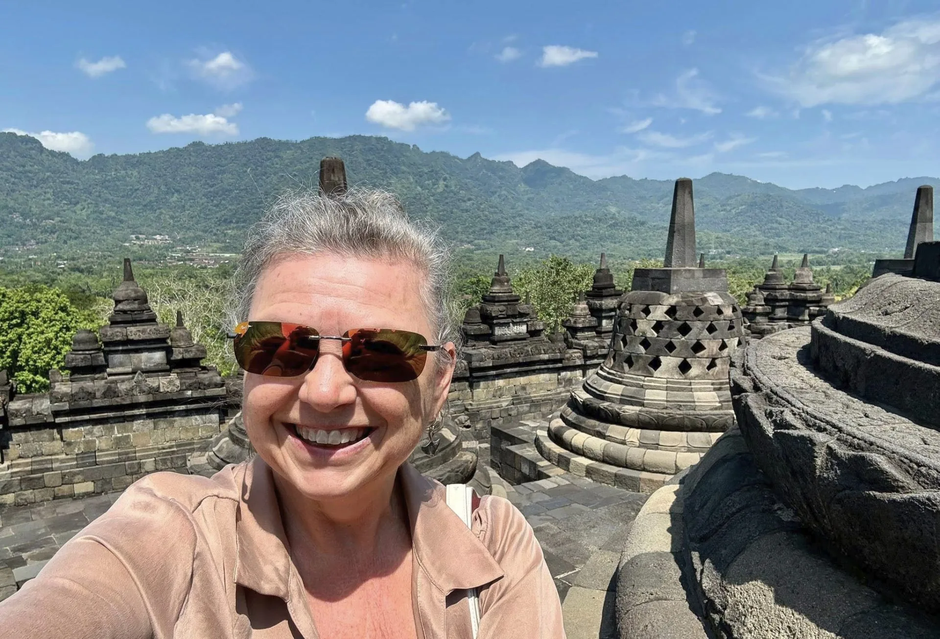 Smiling woman in sunglasses taking a selfie among the bell-shaped stone stupas of Borobudur Buddhist temple with mountains in the background in Central Java, Indonesia.