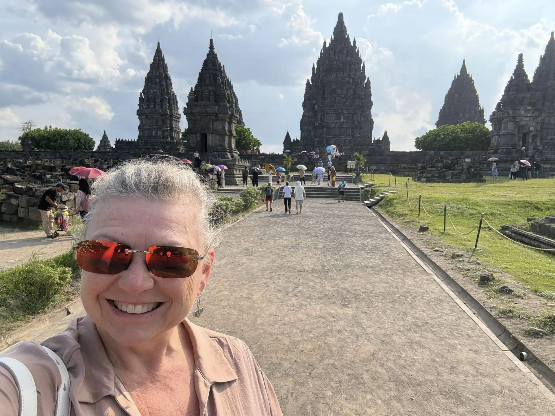 Smiling woman in sunglasses taking a selfie in front of the tall carved spires of the Prambanan Hindu temple complex in Central Java, Indonesia.