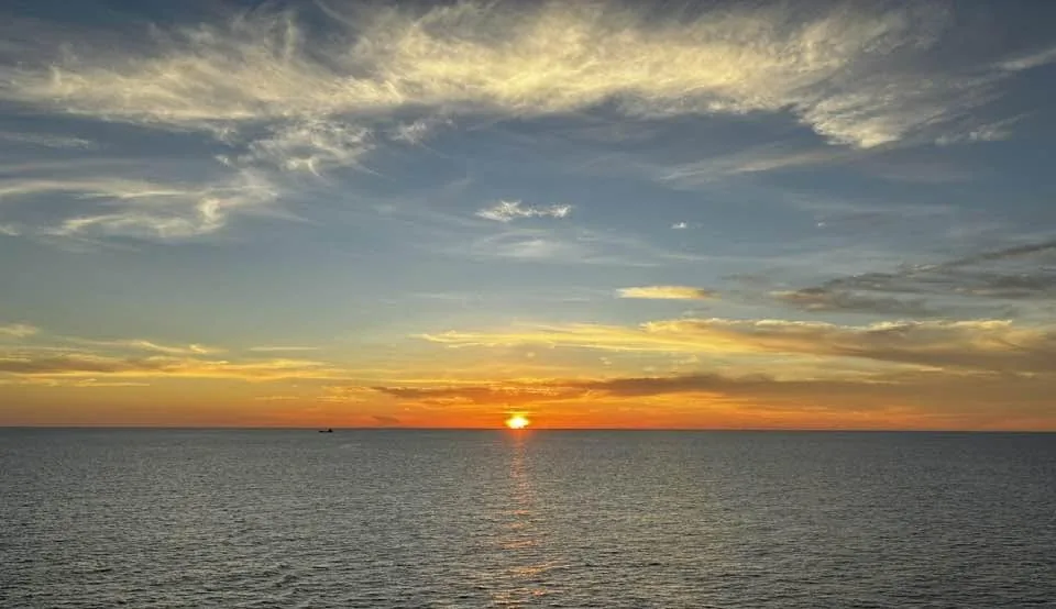 Golden and orange sunset over the calm South China Sea with wispy clouds streaking the sky and a small boat on the horizon.