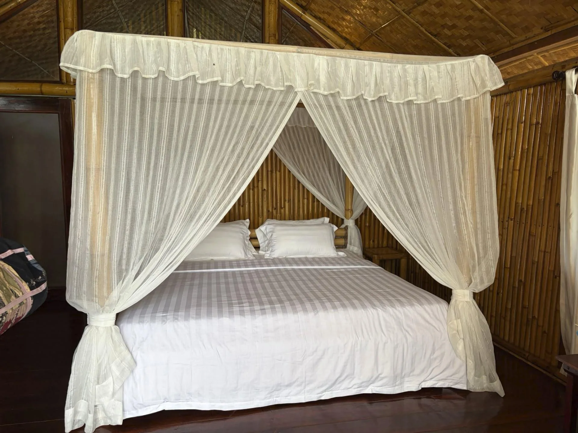 Wooden four-poster bed draped in white sheer mosquito netting inside a bamboo and thatch bungalow with woven walls on Tenggiling Island, Indonesia.
