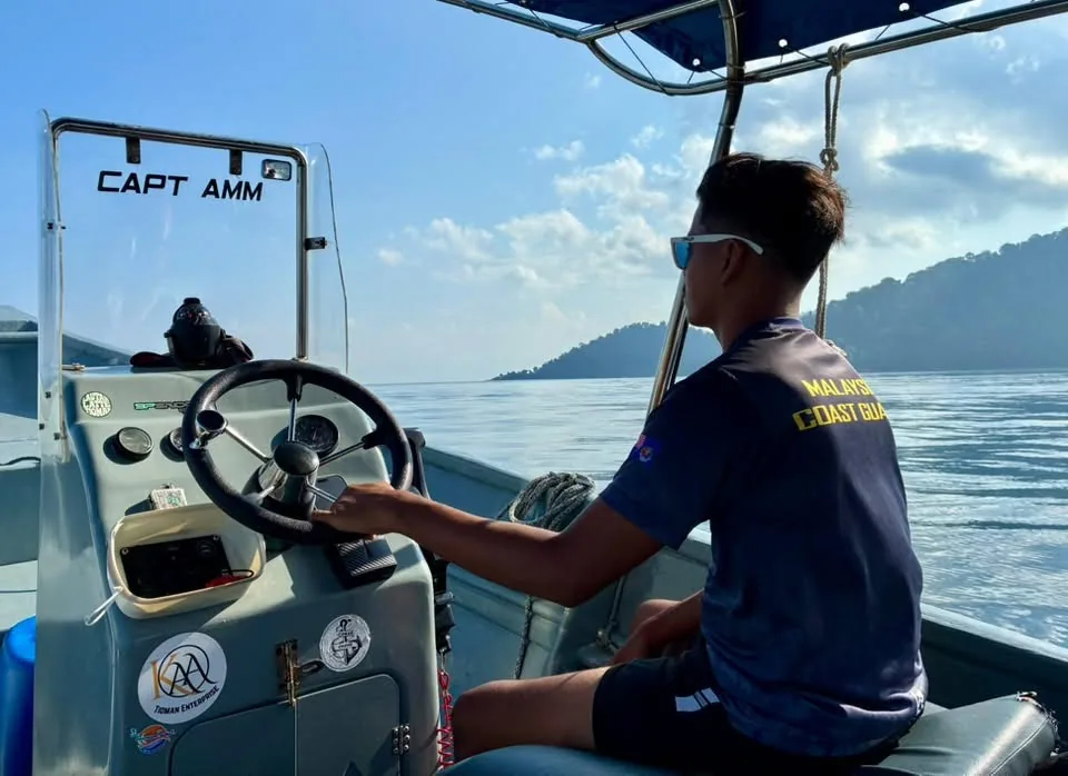 Malaysian Coast Guard crewman in sunglasses steering a small boat named Capt Amm across calm blue water toward a green island near Tioman Island, Malaysia.
