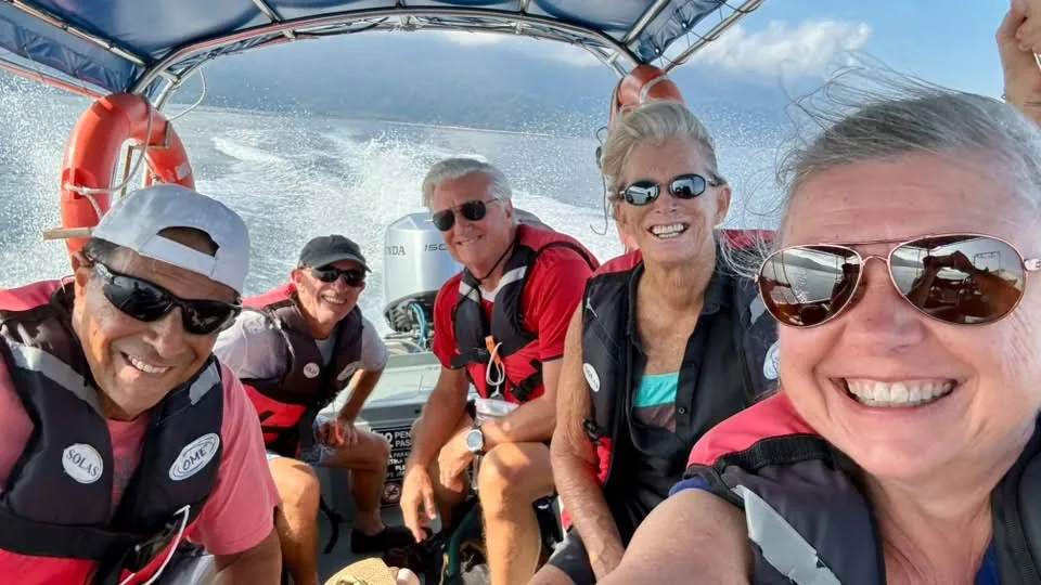 Group of five smiling travelers in red and black life vests taking a selfie on a speedboat leaving a wake across the water near Tioman Island, Malaysia.