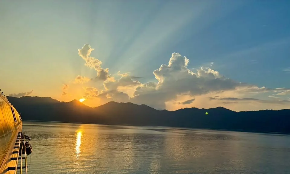 Dramatic sunset with sunbeams radiating behind mountain ridges over calm water, seen from a ship's deck near Tioman Island, Malaysia.